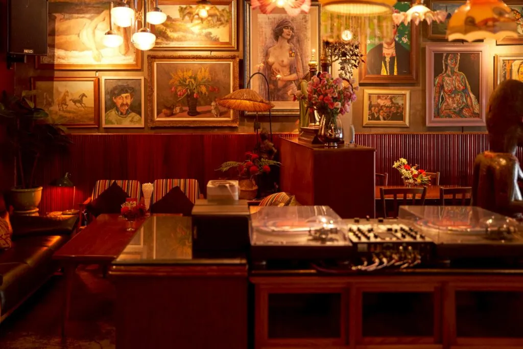 Warmly lit dining room at TUBA Bangkok with antique oil paintings, fringed lamps and a wooden bar counter