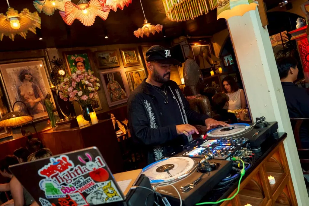 DJ playing vinyl decks at TUBA Bangkok in front of a wall of framed vintage paintings
